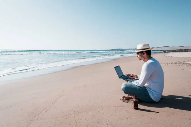 man sitting on the beach with a laptop alone doing telecommuting or remote work and drinking beer