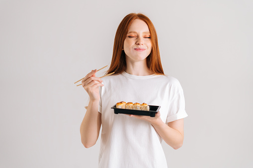 Studio portrait of satisfied young woman with closed eyes eating delicious sushi rolls with chopsticks standing on white isolated background. Happy Caucasian redhead female chewing Asian food.