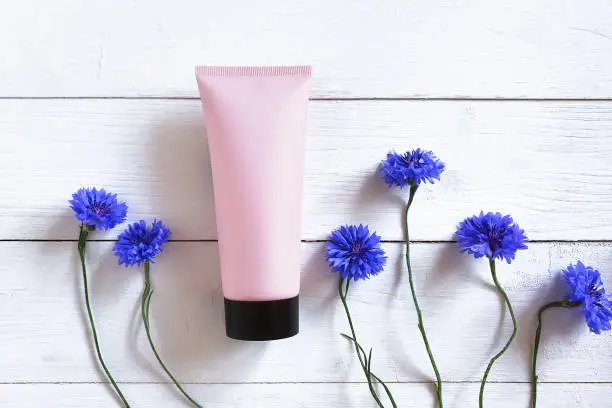 Photo of Top view of mockup light pink squeeze bottle plastic tube with black cap and  blue cornflowers flowers on white wood background. Natural organic spa cosmetics concept. Flatlay, front top view.