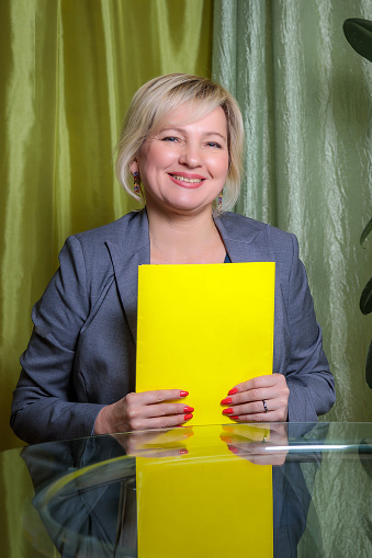 A pretty middle-aged woman, director, sits at a table in the office, holding a yellow folder with documents. Smiling for the camera.