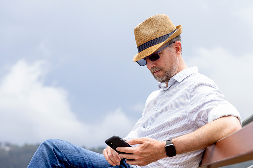 Mature man with straw hat sitting outdoor and using his smartphone, background with copy space, full frame horizontal composition