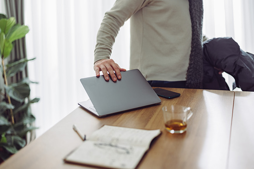 Close up photo of businessman hand closing laptop computer while standing at desk in the living room and with scarf and jacket, ready to go out.