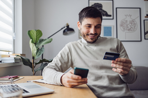 Happy businessman using mobile phone and credit card for online shopping while working from home.