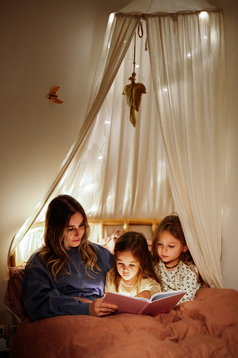 Young Loving Mother Reads Bedtime Stories to Her Little Beautiful Daughters who Goes to Sleep in Her Bed.