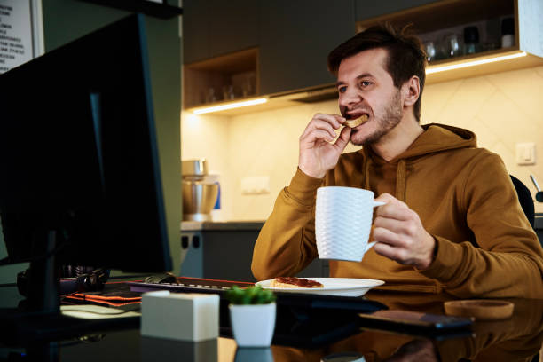 Tired man works late at workplace, use computer Man works at home office workplace eating while works at computer. Freelancer remotely working late. Emotional stress and burnout instant food stock pictures, royalty-free photos & images