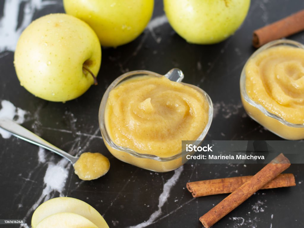 Homemade applesauce (apple puree or mousse) in small glass bowls with organic golden delicious apples and cinnamon sticks on a black textured table Homemade applesauce (apple puree or mousse) in small glass bowls with organic golden delicious apples and cinnamon sticks on a black textured table. Creamy and healthy baby food. Baby Food Stock Photo