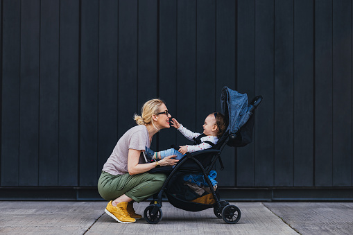Side view of a young Caucasian mother who is playing with a toddler sitting in a stroller. They are outside during the morning or later in the day. They are both wearing casual clothes as if they went outside for a walk. The woman might be a single mother or a babysitter. The small girl looks amused. They are both laughing.