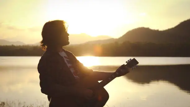 Photo of Man playing acoustic guitar outdoors with sunlight reflected on water surface at sunset lake.