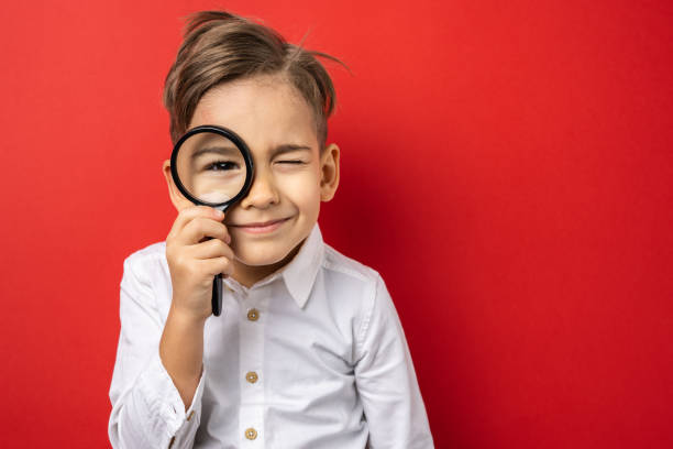 one boy in front of red background wall holding lens magnifying glass looking to the camera copy space front view waist up wearing white shirt smiling curious - magnifying glass lens holding europe imagens e fotografias de stock