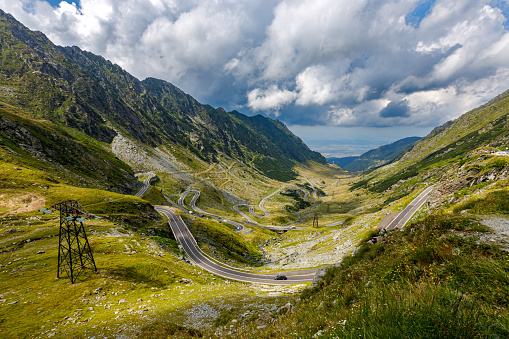 Wide-angle view of the renowned road ascending Passo dello Stelvio (formally known as Strada Statale 38 dello Stelvio), a mountain pass which connects Valtellina and Val Venosta at an elevation of 2,757 metres above sea level, in the Italian Alps, offering breathtaking views on the surrounding mountaintops and the underlying valley. The dazzling bright light of a summer noon, picturesque clouds, steep rocky cliffs and gentle green slopes, cyclists, bikers and cars following the narrow, winding strip of asphalt. High level of detail, natural rendition, realistic feel. Developed from RAW.