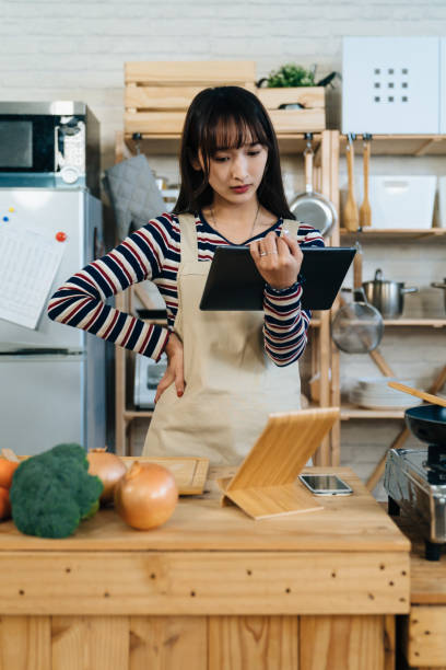 istock vertical shot confused Asian girl standing with tablet computer and arm akimbo is learning cooking by watching online tutorial videos near kitchen table at home 1361686504
