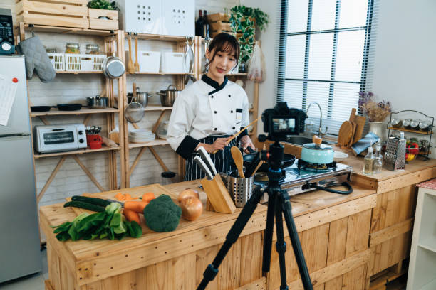 istock Asian woman chef is talking to the digital camera on tripod while filming and teaching cooking online in a bright rustic kitchen at home 1361686801