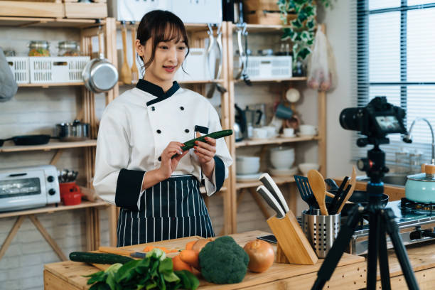 portrait asian female chef recording culinary tutorial video in the kitchen is talking to the camera on tripod while holding and teaching how to choose a cucumber