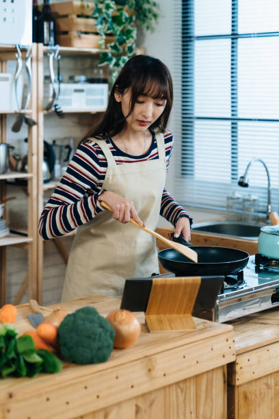 istock vertical portrait of asian new wife is watching online tutorial video on tablet computer to learn cooking lunch while using a frying pan in the kitchen at home. 1361687310