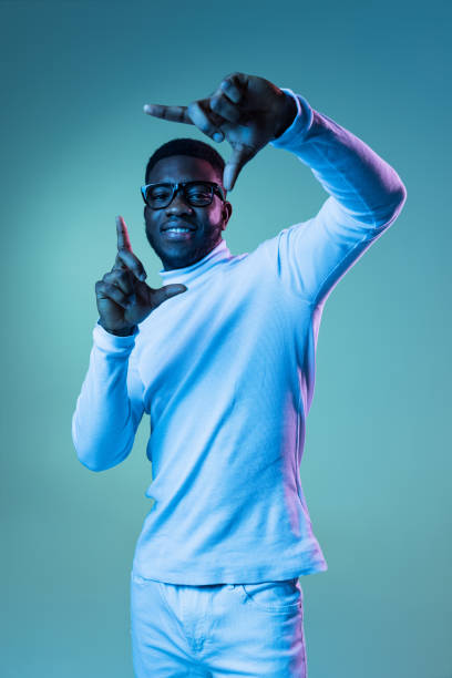 foto de estúdio de um jovem da moda, estudante vestindo pescoço-polo branco e jeans isolados em fundo azul em luz neon. - polo shirt african ethnicity men african descent - fotografias e filmes do acervo