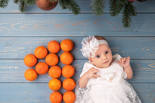 Portrait of a baby girl in a white dress. The baby is one month old. The baby lies on a blue wooden background with a green Christmas tree branch. Number one is made of tangerines.