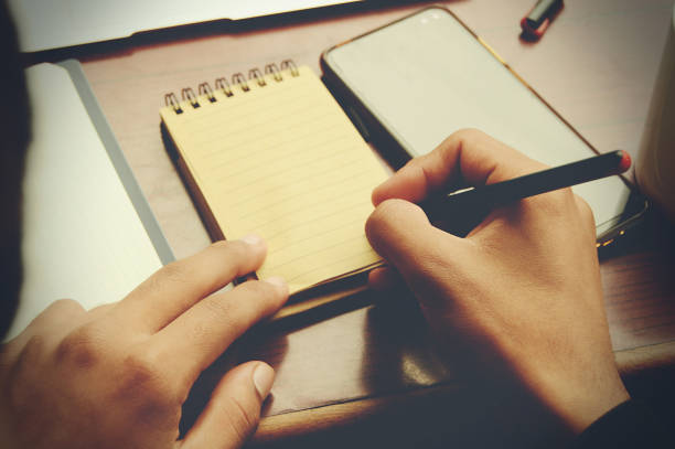 istock Horizontal photograph captured from back of an unrecognisable young man writing on blank page of a spiral notebook,  working on a wooden desk with  mobile phone 1361697935