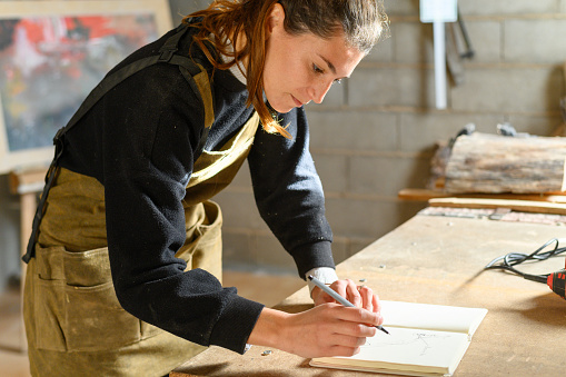 Female artisan in apron leaning on table and writing in notebook during work in sunlit workshop in daytime