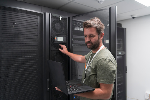 Data Center Engineer Using Laptop Computer. Server Room Specialist Facility with Male System Administrator Working with Data Protection Network for Cyber Security or Cryptocurrency Mining Farm.