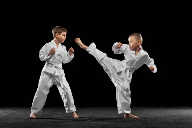 Two little kids, boys, taekwondo athletes training together isolated over dark background. Concept of sport, education, skills, workout, healthy lifestyle and ad. Power and energy.