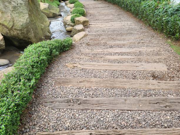 A path with pieces of wood that looks weathered and gravel around it as well as green trees around it is photographed during the day