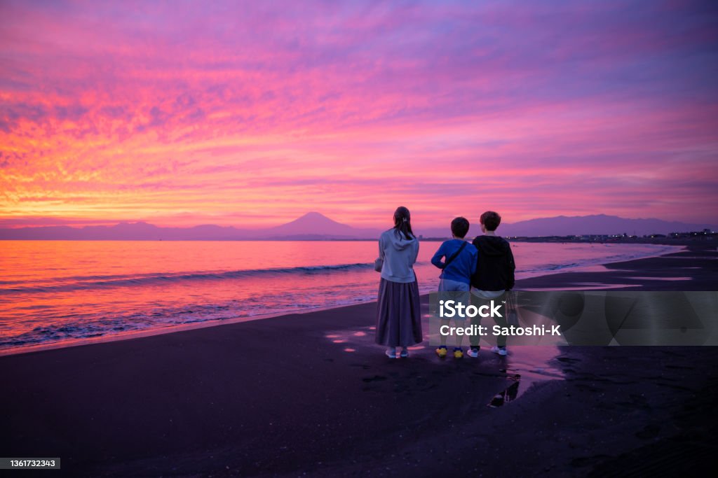Rear view of three people looking at Mt. Fuji from Shonan beach at sunset time Female friends and a son playing badminton and catch at public park, and visiting beach to enjoy beautiful sunset. Pink Color Stock Photo