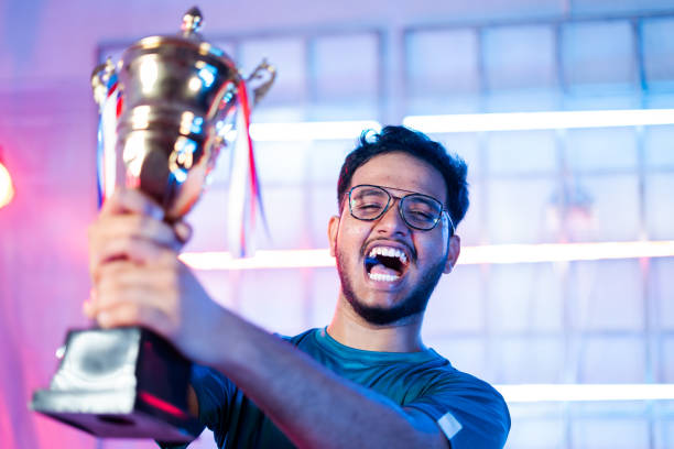 excited gamer celebrating win by showing trophy to audience and shouting by looking at live streaming camera during esports tournament - concept of professional champion gamer. - winners podium audio imagens e fotografias de stock