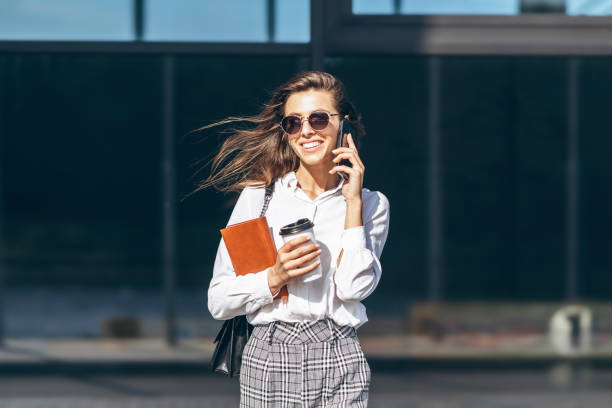 Business woman walking and talking on the cellphone outdoors near modern business center with notebook. stock photo