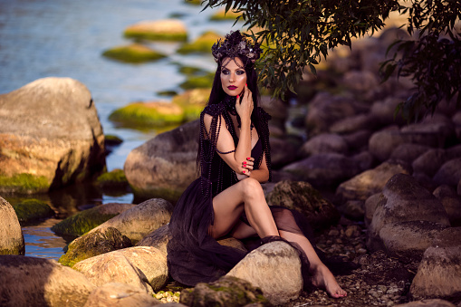 Caucasian Brunette Female Ballerina In Black Dress Posing on Rocky Shore While Wearing Seashell Decorated Crown And Long Black Pearl Necklace. Horizontal Image Orientation