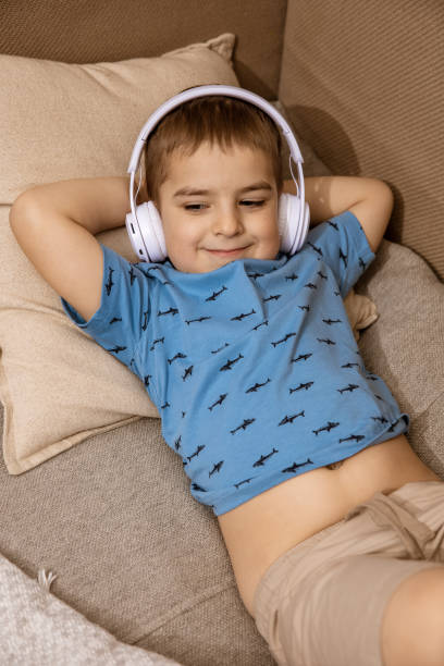 piccolo ragazzo caucasico con camicia blu e cuffie bianche che ascolta musica o audiolibro sul divano di casa. bambino carino che si rilassa, riposa nella sua stanza. - one kid only audio foto e immagini stock