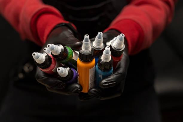 tatuador masculino segurando garrafas de tatuagem de várias cores. foco seletivo nos frascos, fundo preto, vestindo suéter vermelho com luvas pretas. conceito de arte corporal - matéria corante - fotografias e filmes do acervo