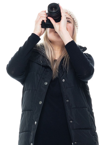 Front view of aged 20-29 years old who is beautiful caucasian female photographer standing in front of white background wearing pants who is laughing who is photographing and holding camera