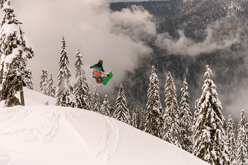 freeride snowboarder performs a masterful jump in the air over a snow-covered mountain slope against a background of snow-covered fir trees
