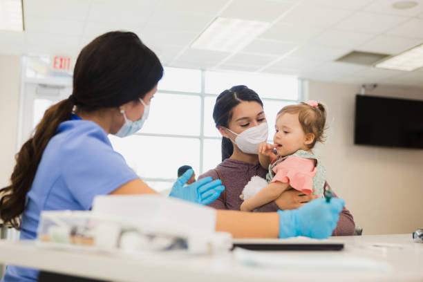 Volunteer nurse cares for baby at free clinic A female healthcare professional gestures as she talks to a young mother and the woman's baby. The nurse and mother are wearing protective face masks during the coronavirus outbreak. emergency medicine stock pictures, royalty-free photos & images