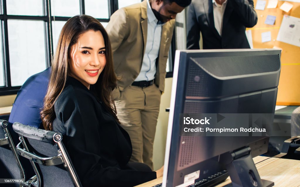 Beautiful smart Asian businesswoman wearing dress, smiling with happiness and success, using laptop for working and networking, looking at camera, sitting on desk in modern indoor office. Business Concept. Beautiful smart Asian businesswoman wearing dress, smiling with happiness and success, using laptop for working and networking, looking at camera, sitting on desk in modern indoor office. Business Concept Adult Stock Photo
