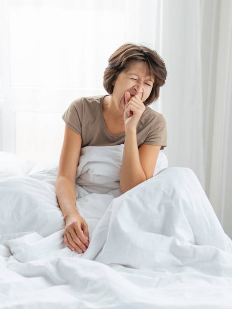 Sleepy woman is yawning in bed, covered with white blanket. It's hard to wake up early in morning. stock photo