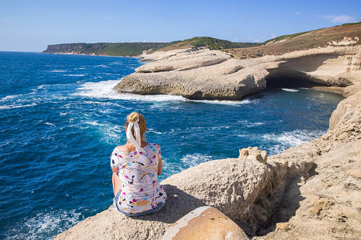 View of a blonde woman sitting alone along the coast near a natural cave, Sardinia, Italy.