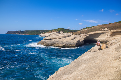 View of a blonde woman sitting alone along the coast near a natural cave, Sardinia, Italy.