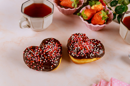 Two heart shaped donuts with white glaze and pink, red sprinkles with strawberry and cup of tea. Valentine Day concept.