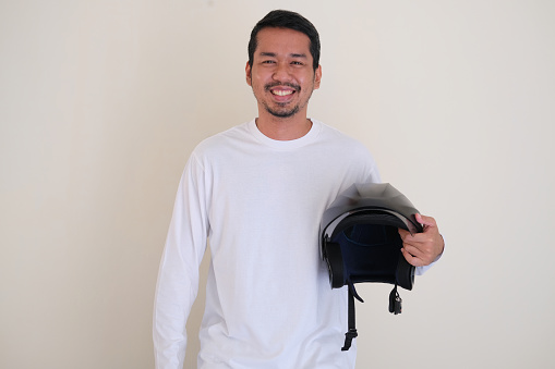 Adult Asian man smiling friendly while holding a motorbike helmet beside