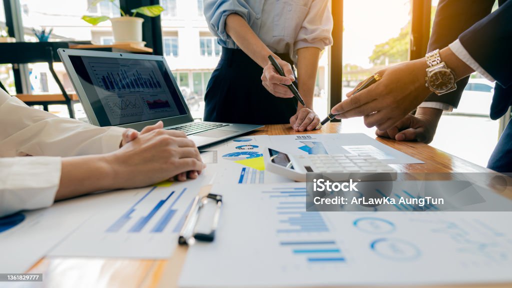Collaborate at office desks to discuss paperwork. A financial group of business people is meeting to analyze data for a marketing plan on a table with graphs and a laptop with a calculator. Teamwork Stock Photo