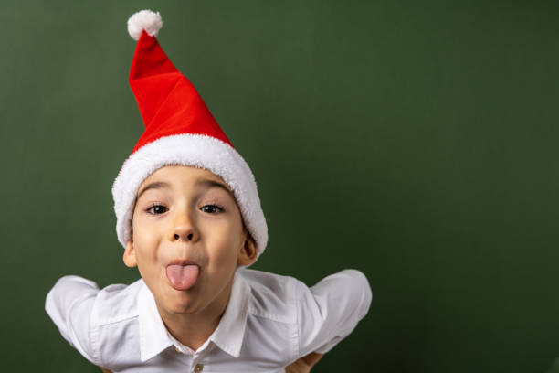 ritratto di un piccolo ragazzo caucasico di cinque anni che indossa un cappello di babbo natale e una camicia bianca di fronte allo sfondo della parete verde che guarda la fotocamera sporgente lingua nuovo anno e natale concetto copy space - surprise child 4 5 years little boys foto e immagini stock