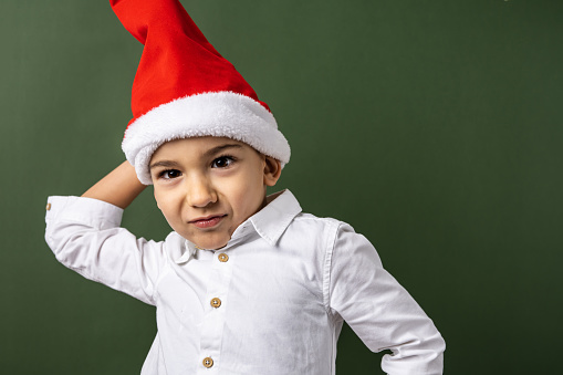 Front view portrait of one small caucasian boy five years old wearing santa hat and white shirt in front of green wall background looking to the camera new year and christmas concept copy space
