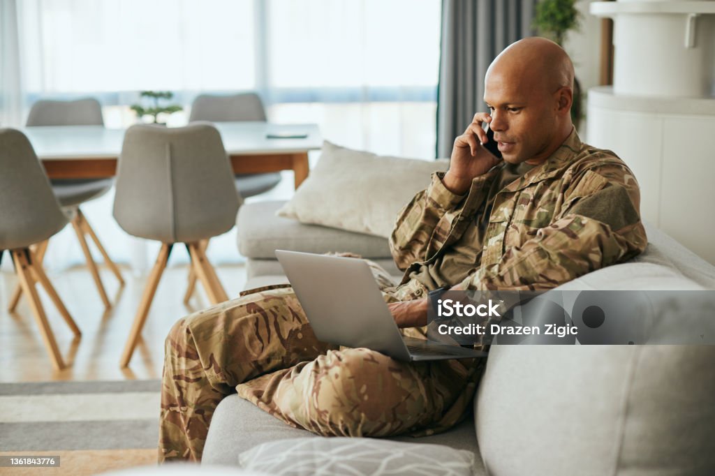 Black military man using laptop while talking on the phone at home. Young African American soldier communicating over cell phone while surfing the net on laptop at home. Veteran Stock Photo