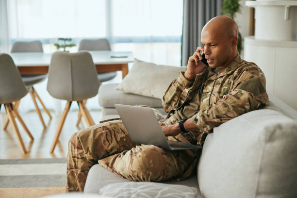 Black military man using laptop while talking on the phone at home. Young African American soldier communicating over cell phone while surfing the net on laptop at home. military lifestyle stock pictures, royalty-free photos & images