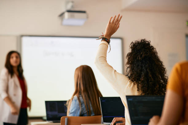 Back view of student raising her hand to ask a question during class in high school. Rear view of high school student raising her arm to ask a question during lecture in the classroom. hand raised classroom student high school student stock pictures, royalty-free photos & images
