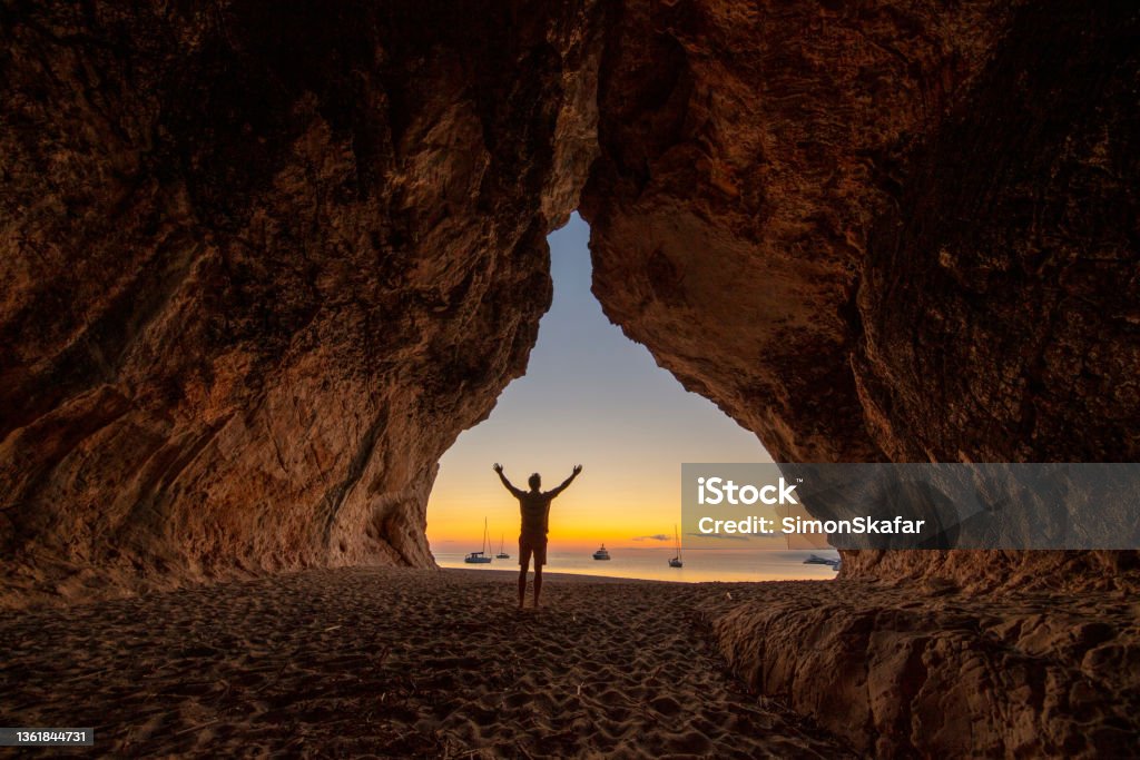 Unrecognizable man at Cala Luna cave at sunset, Gulf of Orosei,Sardinia, Italy. View of a person on the beach at Cala Luna cave at sunset, Sardinia, Italy. Sardinia Stock Photo