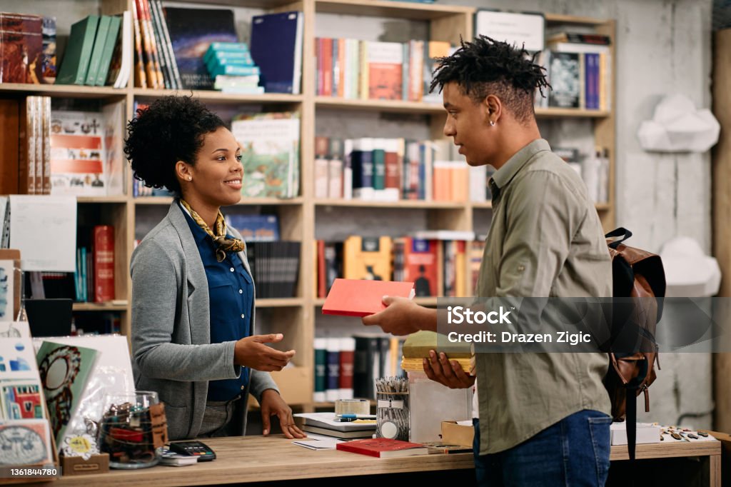 Young black student returning book to a librarian at university library. African American student talking to librarian while returning book at the library. Bookstore Stock Photo