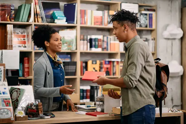 Photo of Young black student returning book to a librarian at university library.