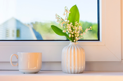 a bouquet of spring lilies of the valley in a small vase stands on the windowsill of the cottage. a cup of morning drink nearby